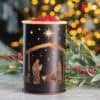 The Nativity Wax Warmer with a nativity scene cutout sits on a marble surface, accented by greenery and red berries, with blurred Christmas lights glowing in the background.