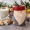 The Gnome Wax Warmer with glowing lights sits on a counter beside a glass of milk, pinecones, decorated gingerbread cookies, and festive greenery in the background.