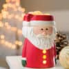 Santa Claus Wax Warmer A Santa Claus Wax Warmer sits on a white surface. In the background, blurred holiday lights, a gold ornament, and a pinecone evoke the cozy spirit of Christmas.