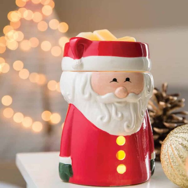 Mint Flip Dish Wax Warmer A Santa Claus Wax Warmer sits on a white surface. In the background, blurred holiday lights, a gold ornament, and a pinecone evoke the cozy spirit of Christmas.