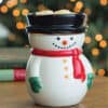 A Frosty Snowman Wax Warmer with a red scarf, green mittens, and black hat sits on a wooden table. The background shows blurred holiday lights and books.