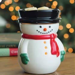 A Frosty Snowman Wax Warmer with a red scarf, green mittens, and black hat sits on a wooden table. The background shows blurred holiday lights and books.