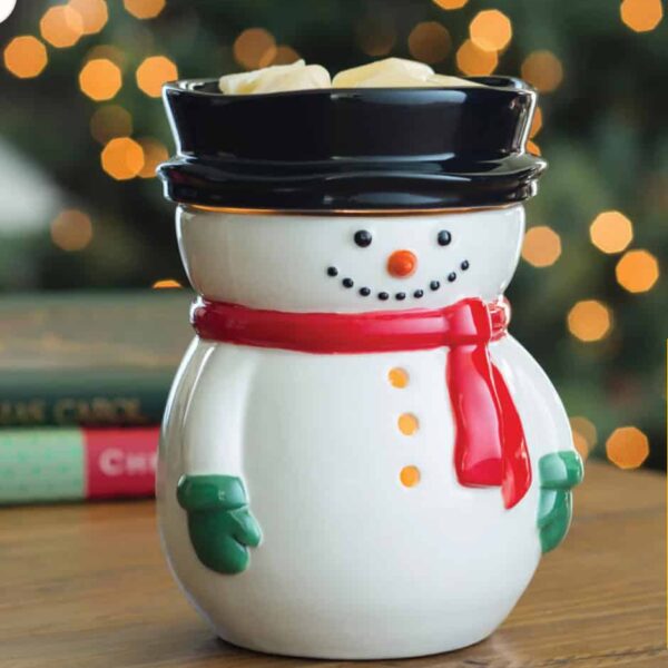 Mint Flip Dish Wax Warmer A Frosty Snowman Wax Warmer with a red scarf, green mittens, and black hat sits on a wooden table. The background shows blurred holiday lights and books.