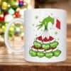 A Personalised Grinch Head Mug features a white Grinch face, green cartoon hand in a Santa sleeve holding up to 7 red and green Santa hats with custom names. A blurred Christmas tree is visible in the background.