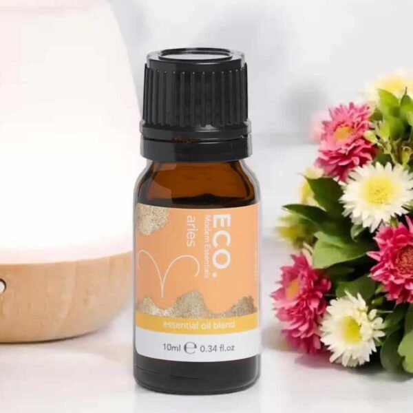 A 10ml amber bottle labeled Aries Zodiac Essential Oil Blend stands on a white surface beside pink and white flowers and part of a wooden diffuser.