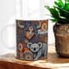 The Taurus Horned Woman with Koala Aboriginal Art Coffee Mug showcases a vibrant design of a woman with long hair, horns, and facial markings holding a koala next to a plant, completed by the word Taurus and its symbol.