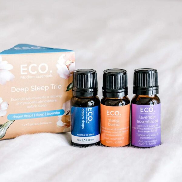 Three small bottles labeled Dream Drops, Sleep Blend, and Lavender rest before a pastel box displaying the product name Deep Sleep Trio Essential Oil 3 Pack, all arranged on a soft white surface.