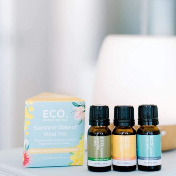 Three ECO. essential oil bottles stand before a pastel Sunshine State of Mind Essential Oil Blends Trio Pack box, with a white diffuser blurred in the background—perfect for uplifting aromatherapy sessions.