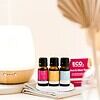 Rest and Glow Essential Oil Trio Pack Three bottles from the Rest and Glow Essential Oil Trio Pack sit on a coaster beside a white diffuser, folded cloth, and pink eco-branded box. Soft lighting adds to the serene atmosphere.