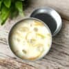 Prosperity & Abundance Travel Tin Candle The Prosperity & Abundance Travel Tin Candle, filled with white wax and adorned with yellow flower petals and translucent stones, sits on a wooden surface near a green leafy plant. Its open lid rests beside the tin.