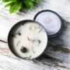 Focus & Achieve Crystal Healing Candle A Focus & Achieve Crystal Healing Candle in a metal tin with crystals and herbs in the wax sits on a rustic white wooden surface, a green plant nearby. The labeled lid rests beside the candle.