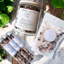 Rose Love Organic Bath Salts are displayed in a jar, pouch, and three glass tubes on a white surface with nearby green leaves. Sunlight casts gentle shadows, emphasizing their elegant presentation.