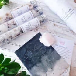 The Little Lux Bath Pack includes three glass bath salt tubes, a white face cloth, a pink aragonite crystal, a dark card, and green leaves arranged on a white wooden surface.