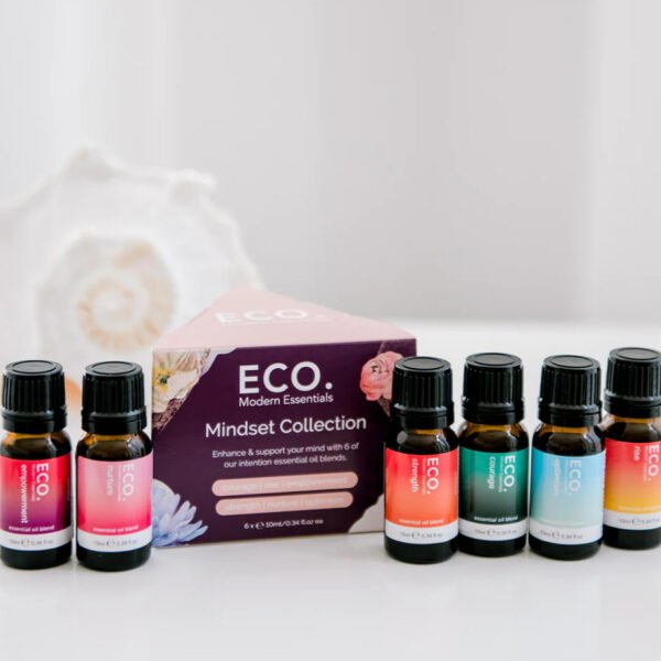 Six bottles of essential oils are arranged in front of a purple Mindset Essential Oil Kit box labeled ECO. The Modern Essentials Mindset Collection is pictured, with a softly blurred decorative white shell in the background.