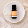 A small amber bottle of Frankincense Pure Essential Oil sits on a white dish, surrounded by pale petals and light fabric on a beige surface.
