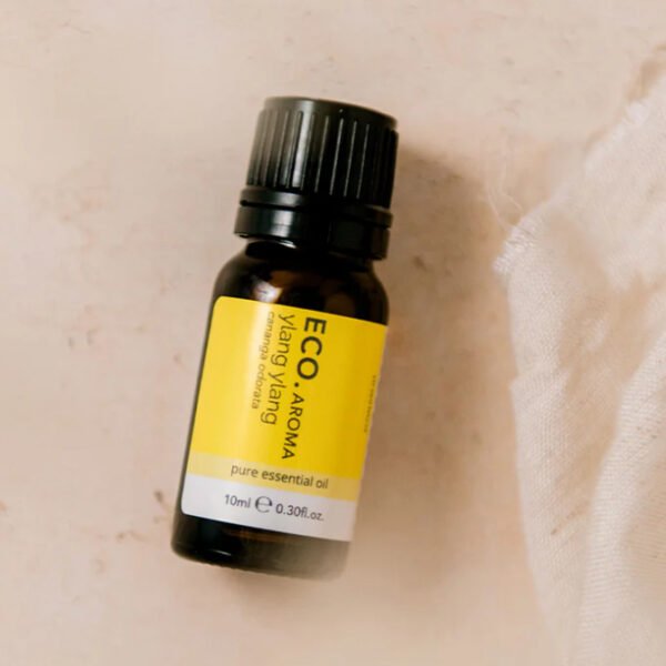Rest and Glow Essential Oil Trio Pack A small dark amber bottle labeled Ylang Ylang Pure Essential Oil sits on a light surface beside a piece of white fabric.
