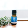 A small brown bottle of Rosemary Pure Essential Oil rests on a white surface beside fresh rosemary sprigs, set against a bright, blurred background.