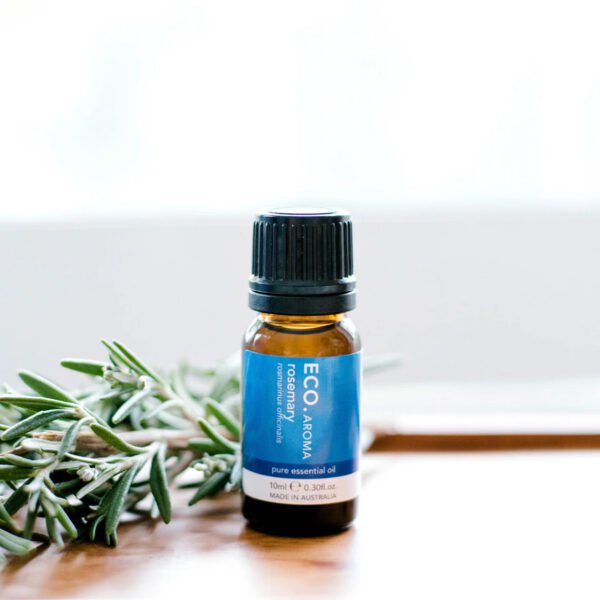 A small brown bottle of Rosemary Pure Essential Oil rests on a white surface beside fresh rosemary sprigs, set against a bright, blurred background.