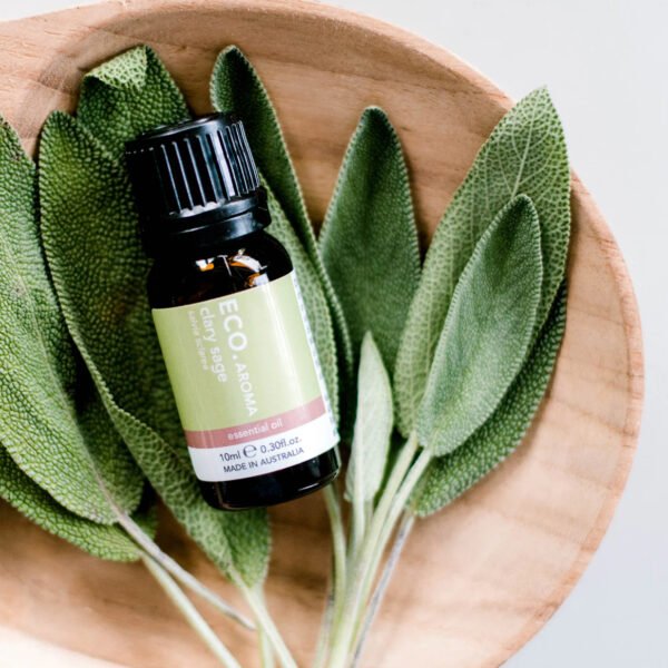 A bottle of Clary Sage Pure Essential Oil sits on fresh sage leaves in a wooden bowl, viewed from above on a light background.