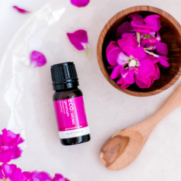 A bottle of Geranium Pure Essential Oil rests on a white surface beside a wooden bowl of pink flower petals and a wooden spoon, with scattered petals adding a vibrant touch to the scene.