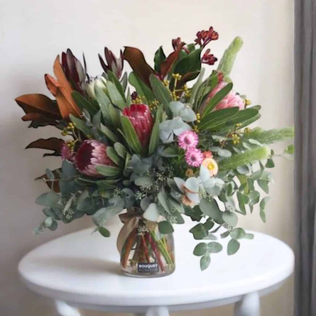 Your Guide to Virgo Season 2025: Symbols, Gifts & Crystals 4 A bouquet of assorted native flowers and greenery in a glass vase sits on a small white round table against a light-coloured wall, perfect for celebrating Virgo Season 2025 or pairing with your favourite Virgo crystals.
