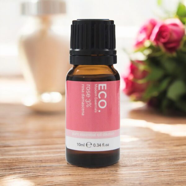 A small amber glass bottle labeled Rose Essential Oil 3% rests on a wooden surface, with a blurred vase and pink roses softly framing the background.