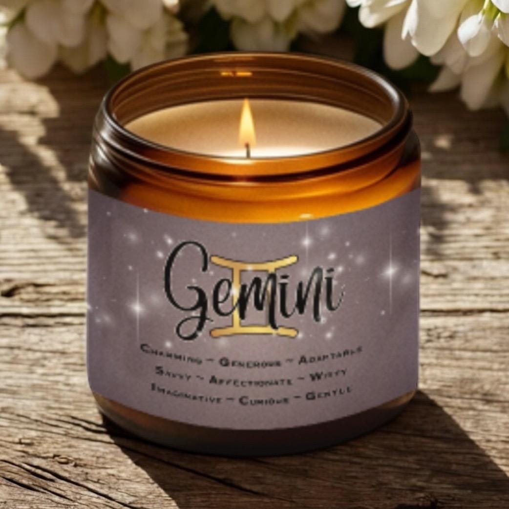 A lit brown glass candle with a label reading Gemini and descriptive traits, placed on a rustic wooden surface with white flowers in the background.