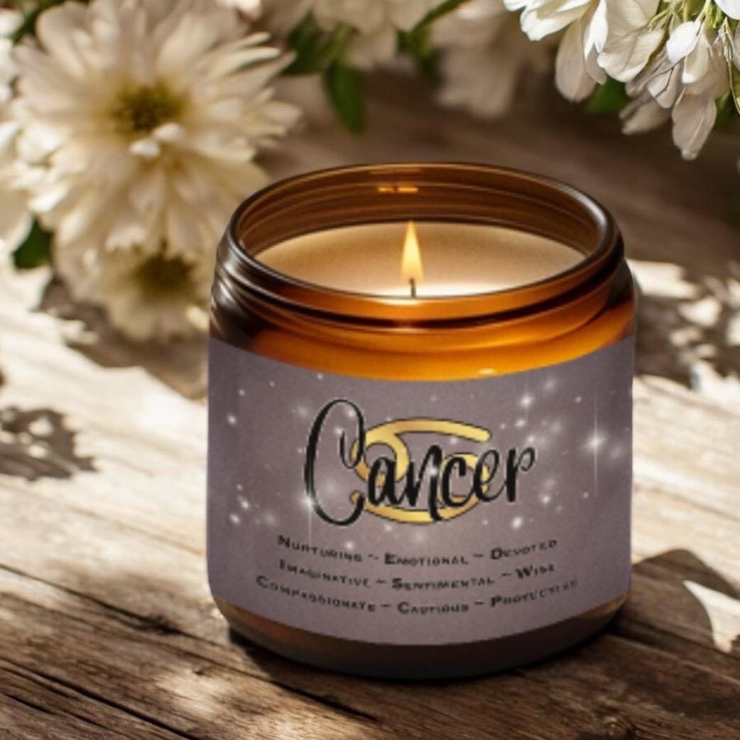 A lit brown glass candle labeled Cancer with zodiac traits sits on a wooden surface beside white flowers and green leaves, illuminated by sunlight.