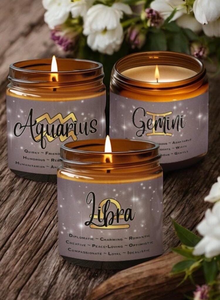Three lit candles in amber jars, each labeled with a zodiac sign—Aquarius, Gemini, and Libra—along with descriptive traits. Flowers are blurred in the background, creating a cozy and calming atmosphere.