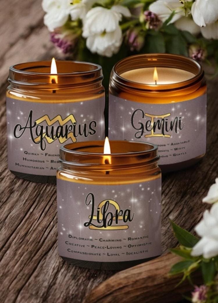 Three lit candles in amber jars, each labeled with a zodiac sign—Aquarius, Gemini, and Libra—along with descriptive traits. Flowers are blurred in the background, creating a cozy and calming atmosphere.