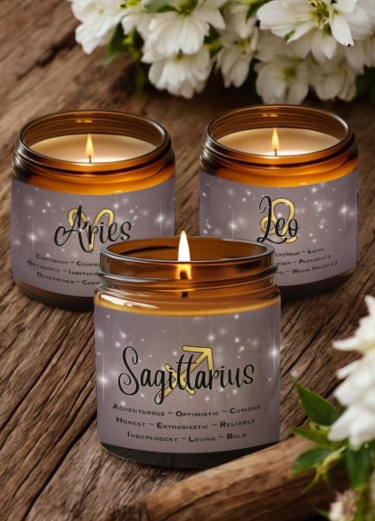 Three brown glass candles are labeled with zodiac signs: Aries, Leo, and Sagittarius. Each candle lists personality traits beneath the sign name. The background features white flowers and a rustic wooden surface.