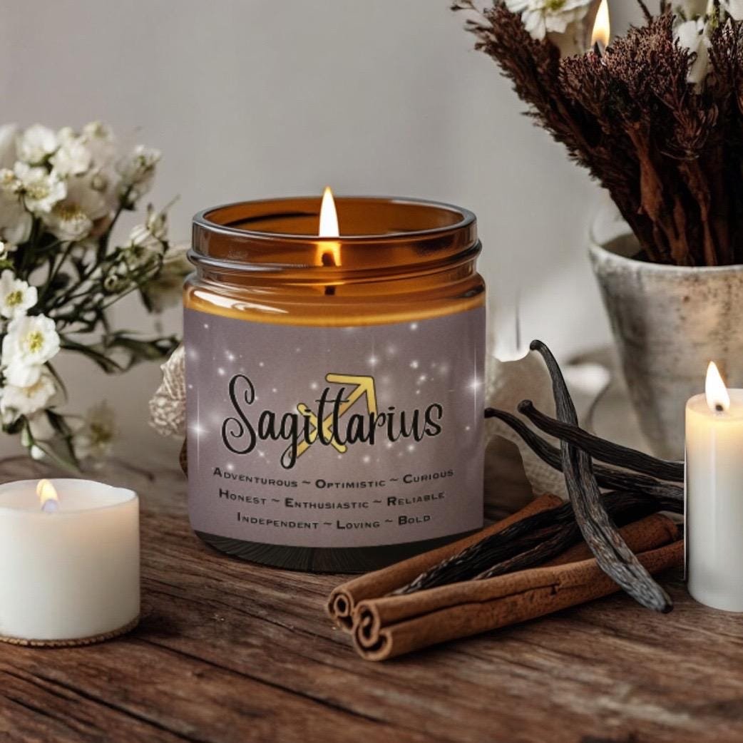 A Sagittarius-themed candle sits on a table with cinnamon sticks, vanilla pods, white flowers, and lit candles. The candle label lists traits like adventurous, optimistic, and honest, with a constellation design in the background.