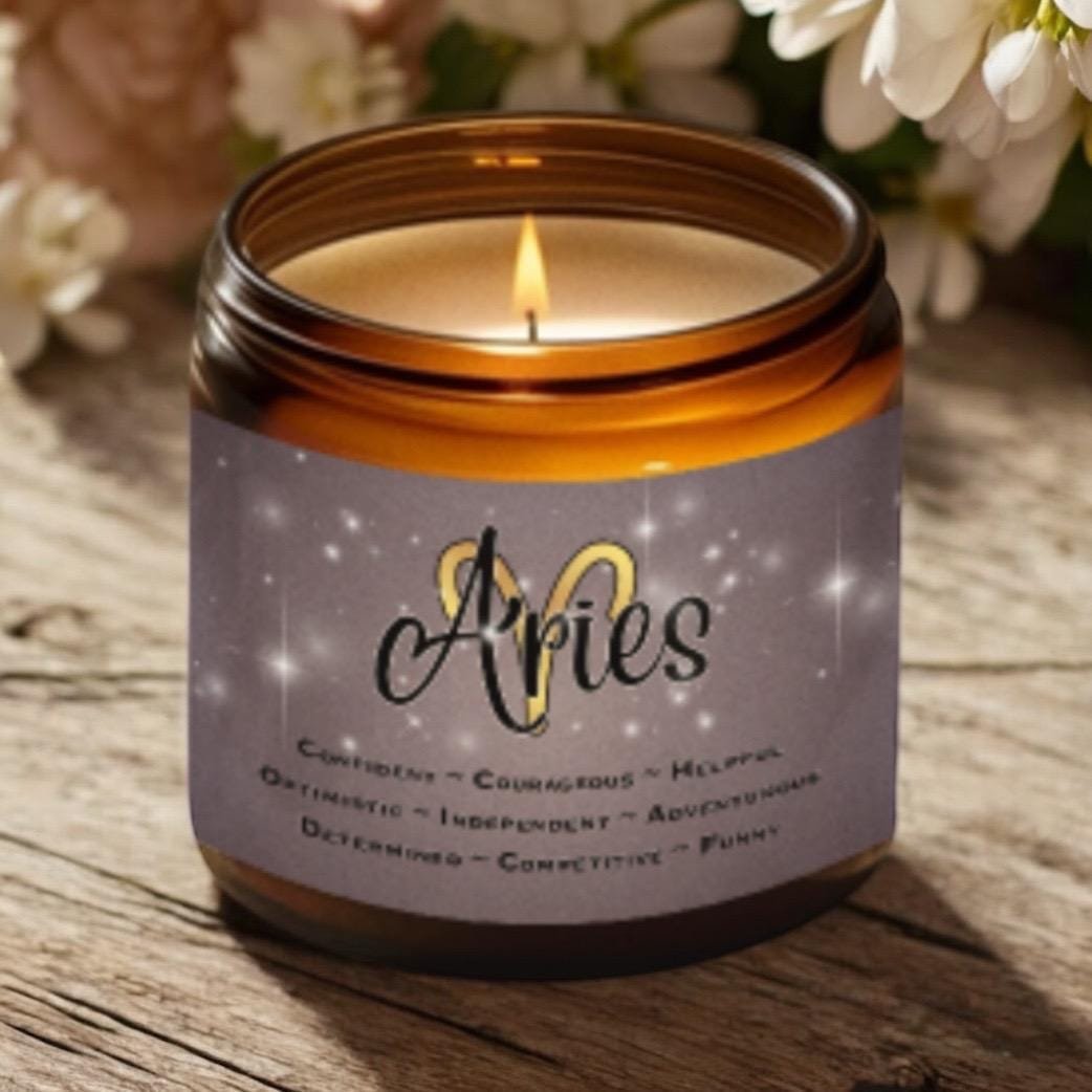 A lit brown glass candle sits on a wooden surface with flowers in the background. The label reads “Aries” and lists traits: confident, courageous, helpful, optimistic, independent, adventurous, determined, competitive, funny.