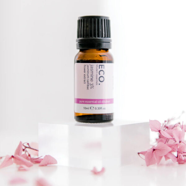 A small brown bottle of Jasmine Essential Oil rests on a clear acrylic block, surrounded by pink dried flower petals against a white background.