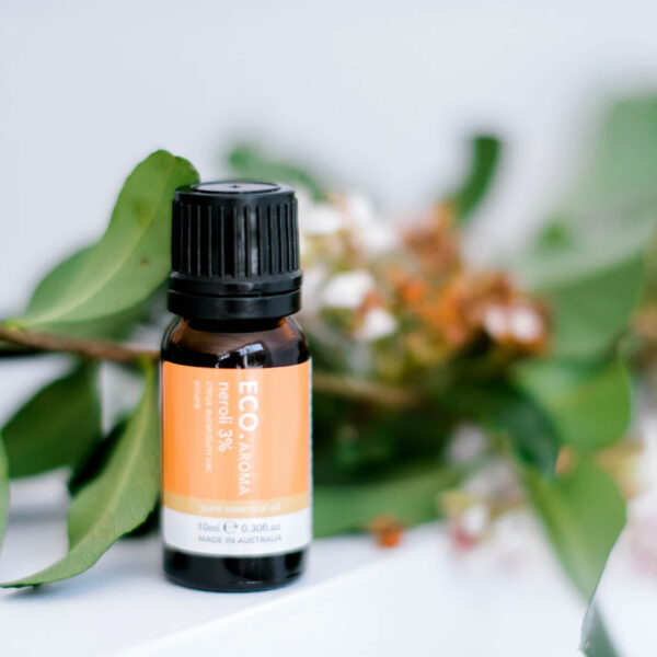 A small amber glass bottle of Neroli Essential Oil 3% in Jojoba stands upright on a white surface, with blurred green eucalyptus leaves and small white flowers in the background.