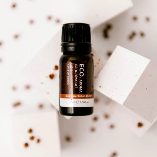 A small dark bottle labeled Sandalwood Essential Oil 10% rests on white geometric blocks, surrounded by scattered brown specks.