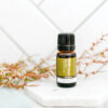 A small amber bottle of Tea Tree Essential Oil – 100% Pure Australian Melaleuca Alternifolia sits on a white marble surface, with delicate greenery in the background and a white tiled wall behind.