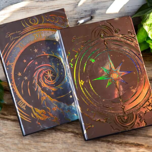 Two Cosmic Clock A5 Hardcover Notebooks with metallic, holographic covers and celestial designs rest on a wooden surface amid green leaves—ideal as astrology journals or for taking cosmic-themed notes.
