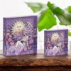 Two decorative blocks with a Virgo design sit on a wooden surface. The design features a woman with flowing hair, a sun behind her, and purple flowers. Green plant leaves are blurred in the background.